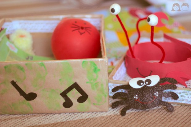 Na imagem #paracegover: Caixa musical aberta, com um pintinho amarelinho, um balão vermelho com um gatinho desenhado, uma aranha de eva brilhante e o chapéu de caranguejo que eu fiz pro Toni.