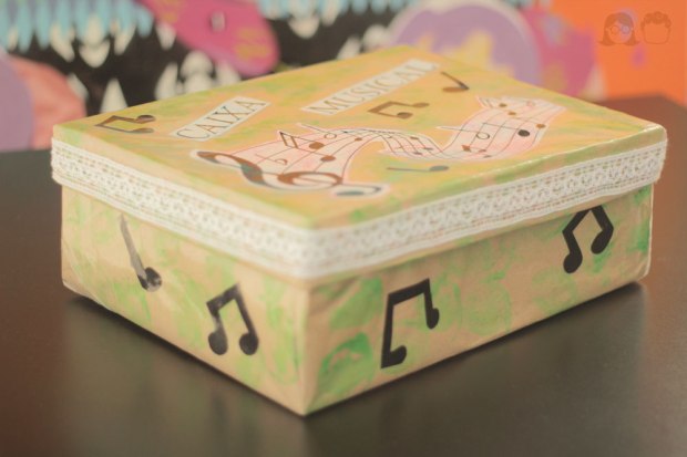 Na imagem #paracegover: Caixa forrada de papel craft, decorada com notas musicais pretas. Na tampa está escrito "Caixa Musical".